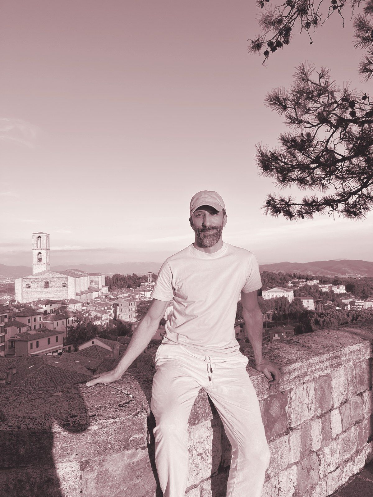 Walter sitting on an old stone wall at golden hour, a hill town and its bell tower behind him.